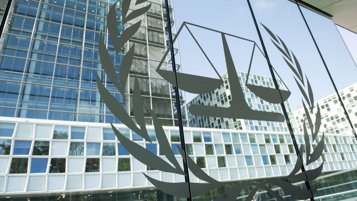 Logo des Internationalen Strafgerichtshof auf einer Fensterfront, eine Waage umkreist von einem oben offenen Lorbeerkranz. Dahinter Gebäude.