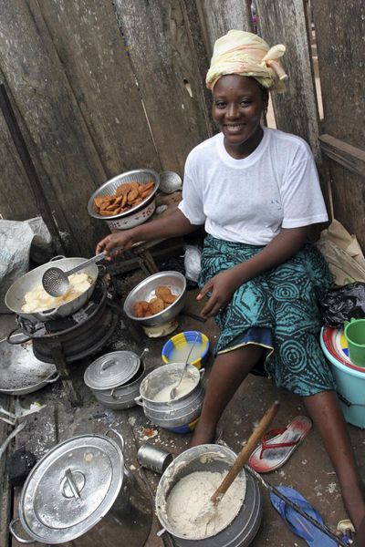 Eine afrikanische Frau sitzt vor einer Bretterwand. Vor und neben ihr stehen Blechschüsseln und -töpfe mit Essen.