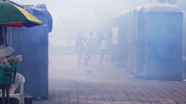 Dichte Schwaden an Tränengas verhüllen den Blick auf die Szenerie. Im Hintergrund fliehen drei Menschen vor dem Gas zwischen blauen Wellblechhütten.