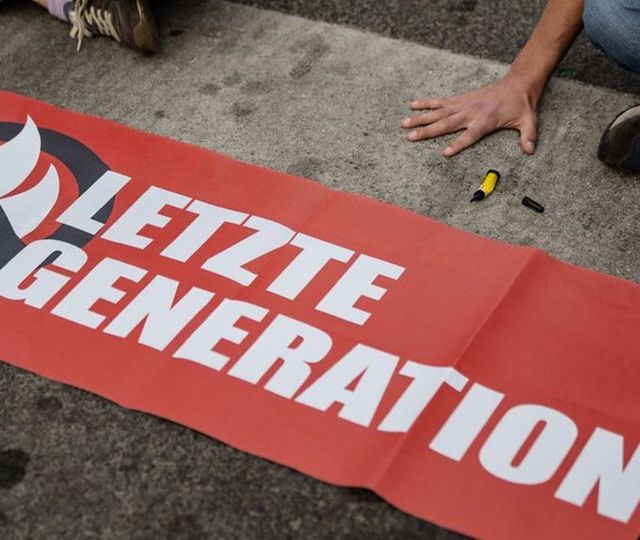 Ein Banner mit der Aufschrift Letzte Generation liegt auf einer Straße, oberhalb sieht man eine festgeklebte Hand.