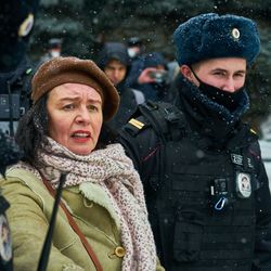 Zwei Männer mit Mundschutz und eine Frau in grünem Mantel auf einem grauen Platz.