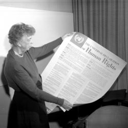 Eleanor Roosevelt mit einem Exemplar der Allgemeinen Erklärung der Menschenrechte im Jahr 1949. Das schwarz-weiß-Foto zeigt eine Frau mit lockigen Haaren, die ein großes Blatt Papier hochhält, auf dem "The Universal Declaration of Human Rights" steht.