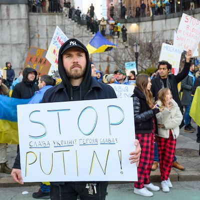 Im Vordergrund steht ein Mann mit einem Poster, auf dem "Stop Putin" steht, im Hintergrund sieht man Demonstrantinnen mit der ukrainischen Fahne.