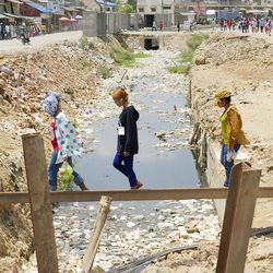 Frauen überqueren einen schmalen, verschutzen Kanal über einen Stahlträger als Brücke