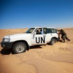 UN Photo/Marine Perret Ein Patroullienfahrzeug der MINURSO steckt im Sand in der Wüste fest, zwei Männer in Uniformen schieben das Auto an.