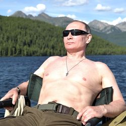 Wladimir Putin sitzt mit nacktem Oberkörper und Sonnenbrille entspannt zurückgelehnt vor einem See.