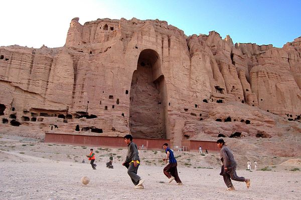 Im Vordergrund spielen Kinder Fußball, im Hintergrund sieht man eine sandfarbene Felswand, in der eine große Aushöhlung in Form eines Buddhas klafft.